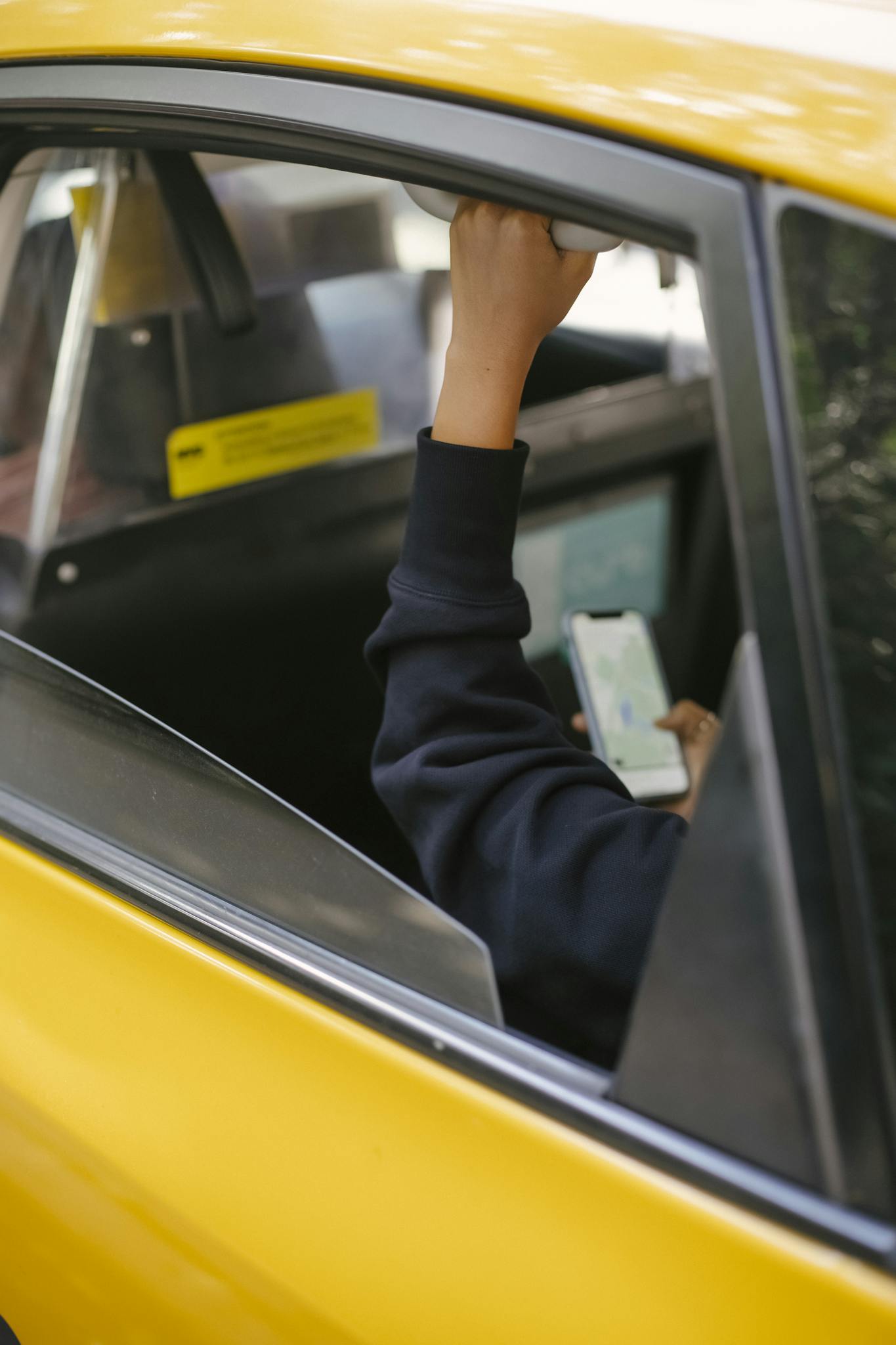 Unrecognizable passenger sitting on backseat and browsing smartphone while holding handle inside of yellow cab with lowered window during ride